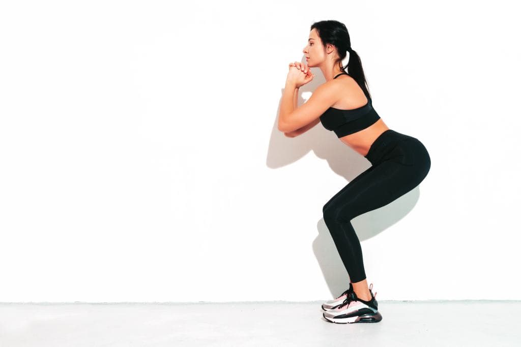 Portrait of fitness confident woman in black sports clothing. Sexy young beautiful model with perfect body. Female isolated on white wall in studio. Stretching out before training. Making squats